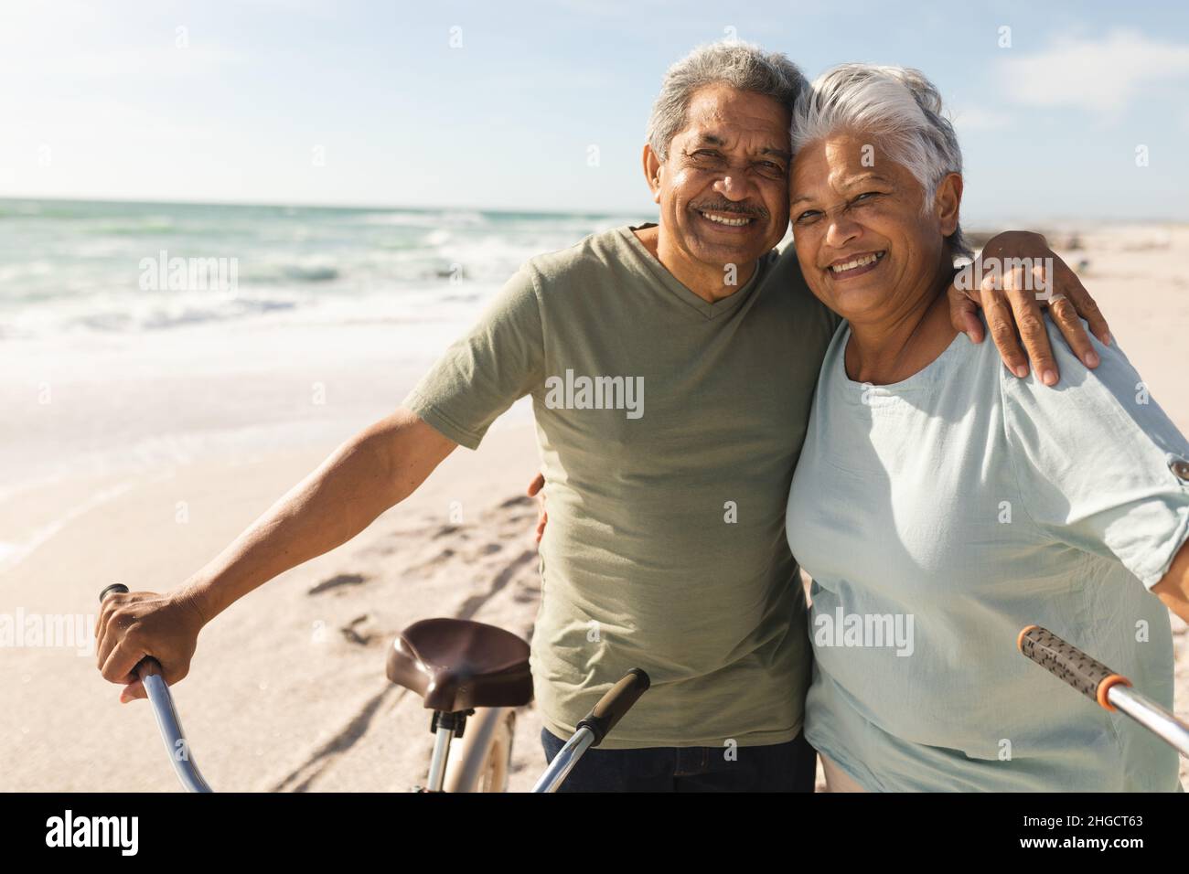 Porträt eines lächelnden mehrrassischen Senioren-Paares mit Arm um Fahrräder am Strand an einem sonnigen Tag Stockfoto