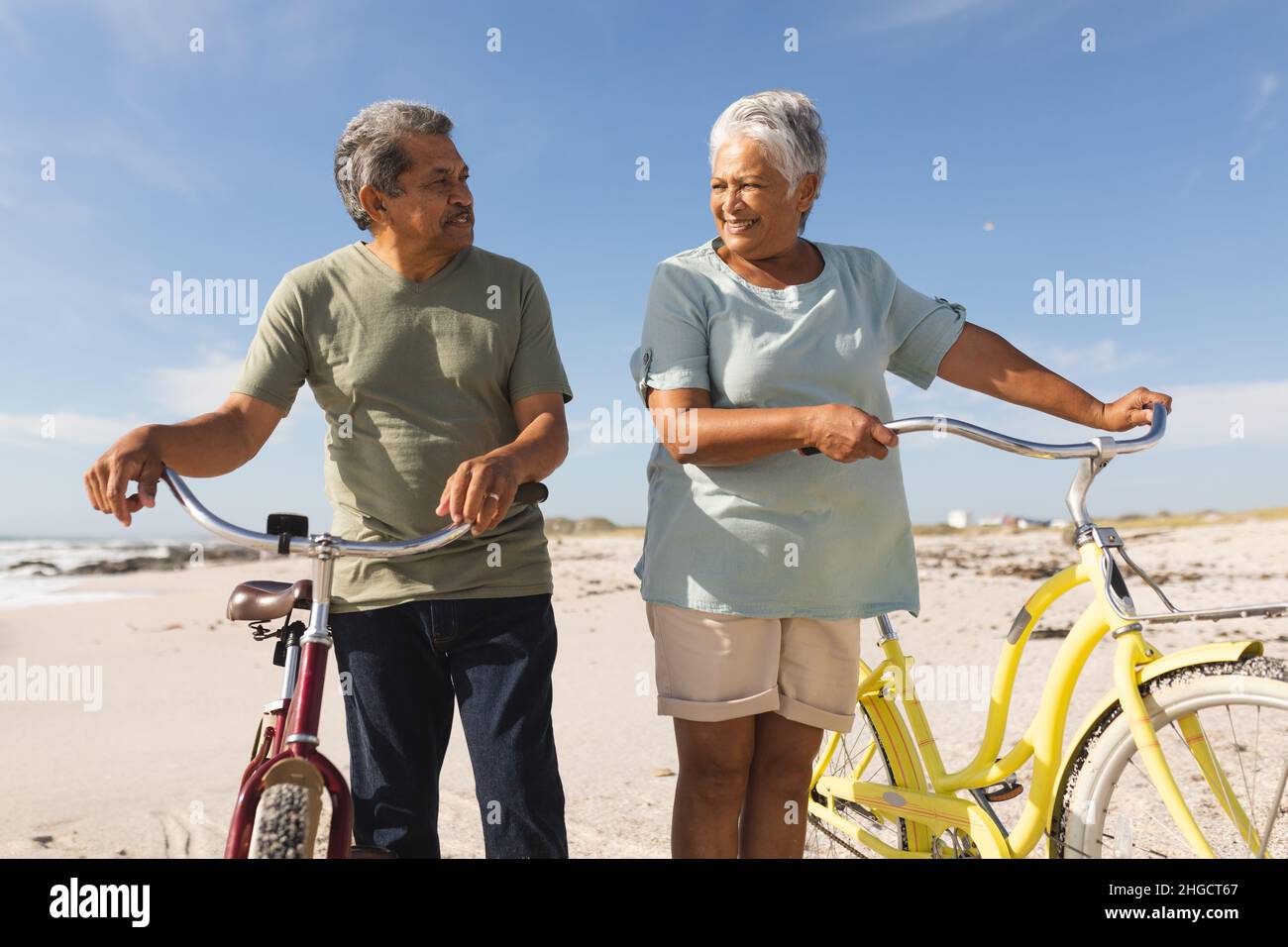 Multirassisches Seniorenpaar, das sich mit Fahrrädern am sonnigen Strand gegen den Himmel ansieht Stockfoto