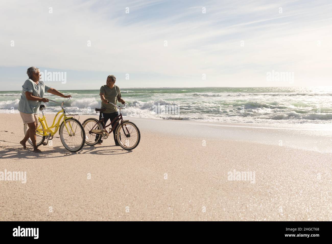 Ältere multirassische Paare reden, während sie am sonnigen Strand gegen den Himmel Fahrräder am Ufer Rollen Stockfoto