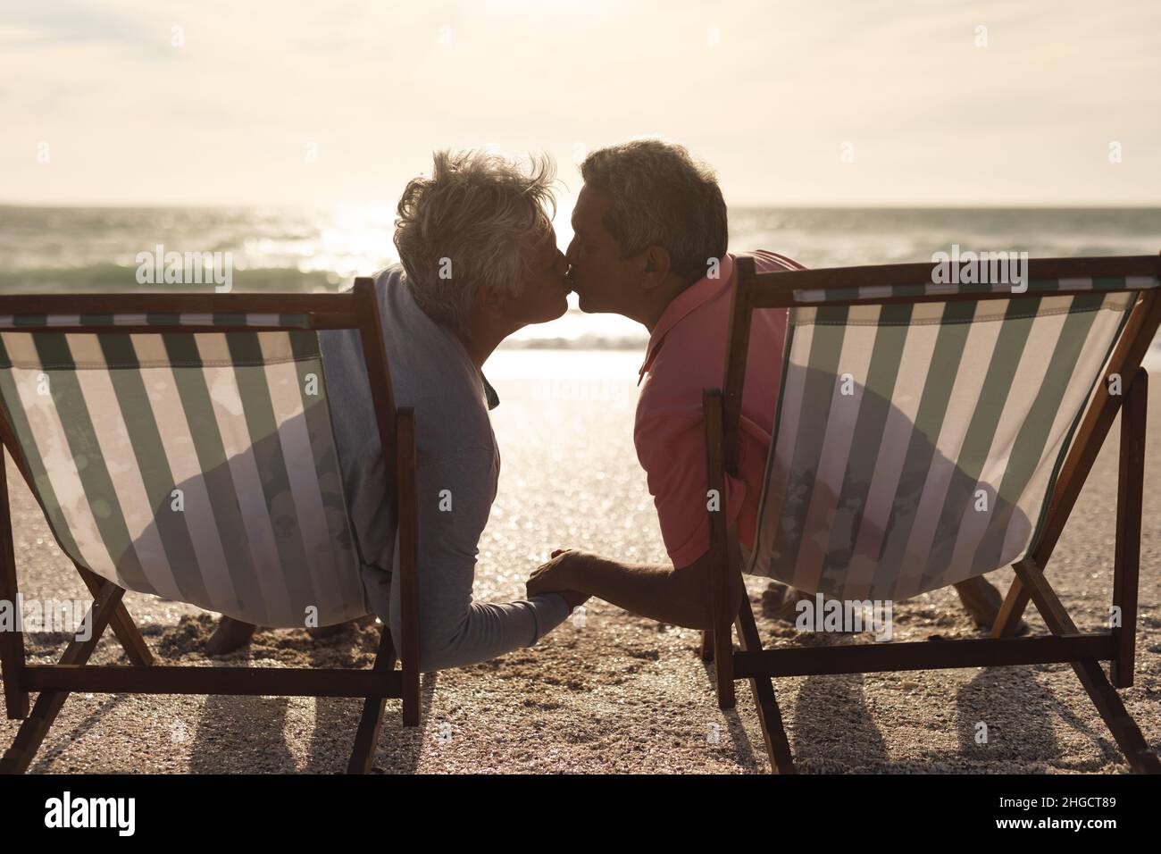 Rentnerpaar mit mehreren Rassen, das auf Klappstühlen sitzt und sich am Strand küsst Stockfoto