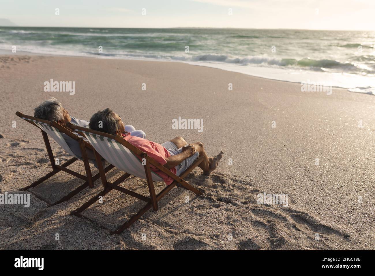 Rentnerpaar mit mehreren Rassen, das sich während des Sonnenuntergangs auf Klappstühlen am Strand entspannt Stockfoto