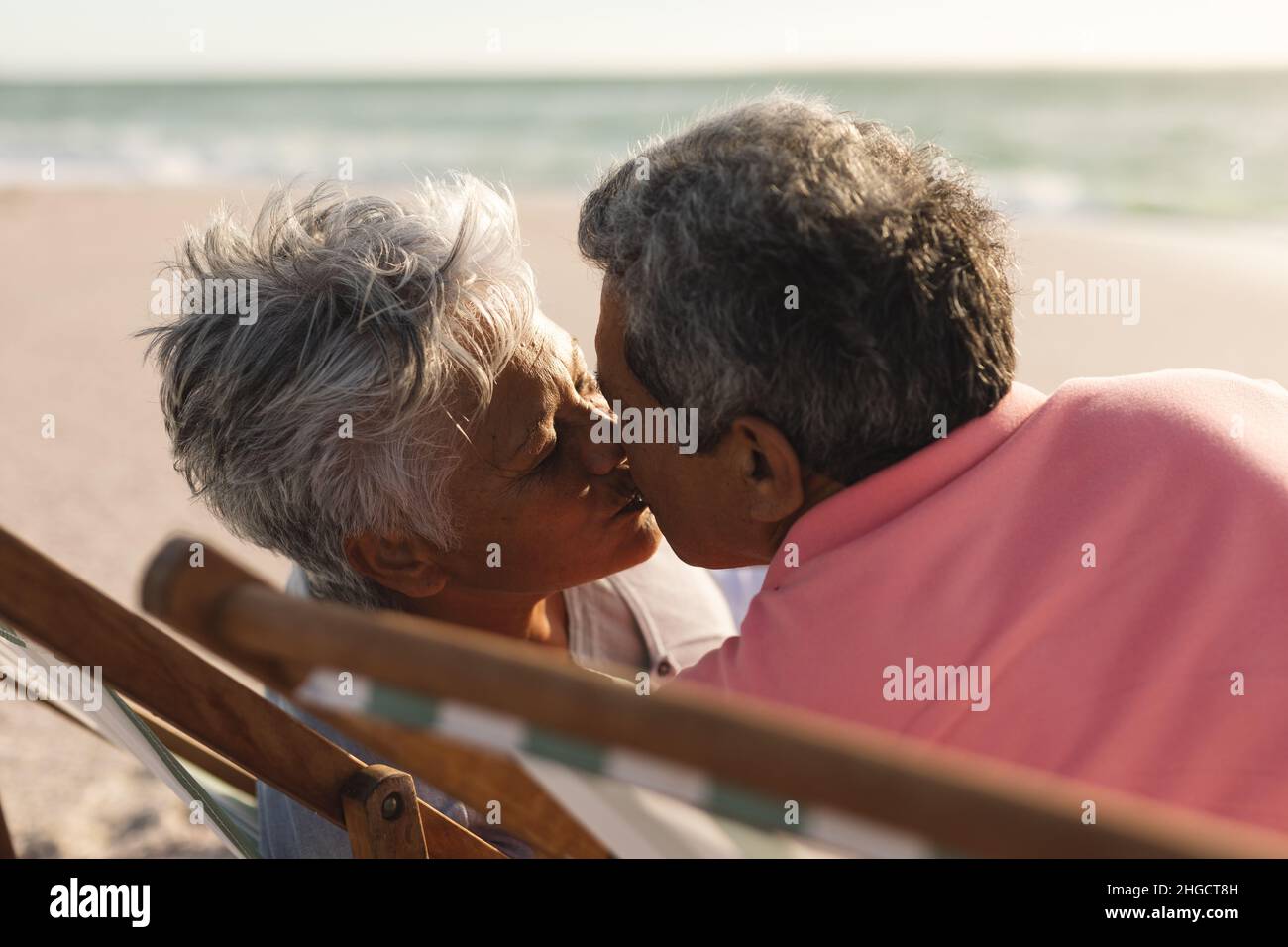 Liebevolles mehrrassisches Seniorenpaar küsst sich, während es am Strand auf Klappstühlen sitzt Stockfoto