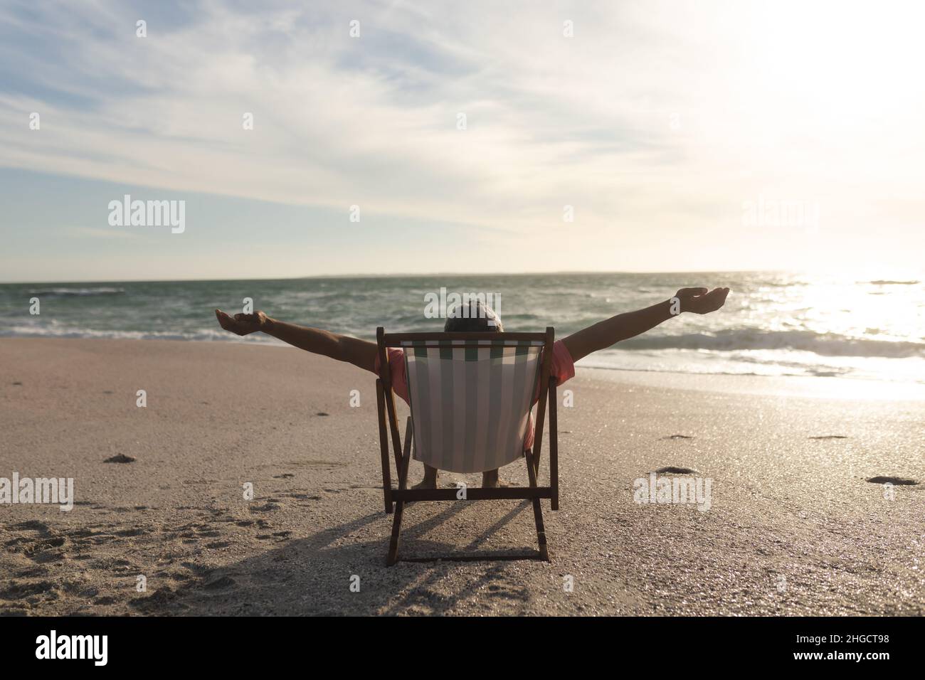Pensionierter älterer Birazialmann mit ausgestreckten Armen, der während des Sonnenuntergangs auf einem Klappstuhl am Strand sitzt Stockfoto