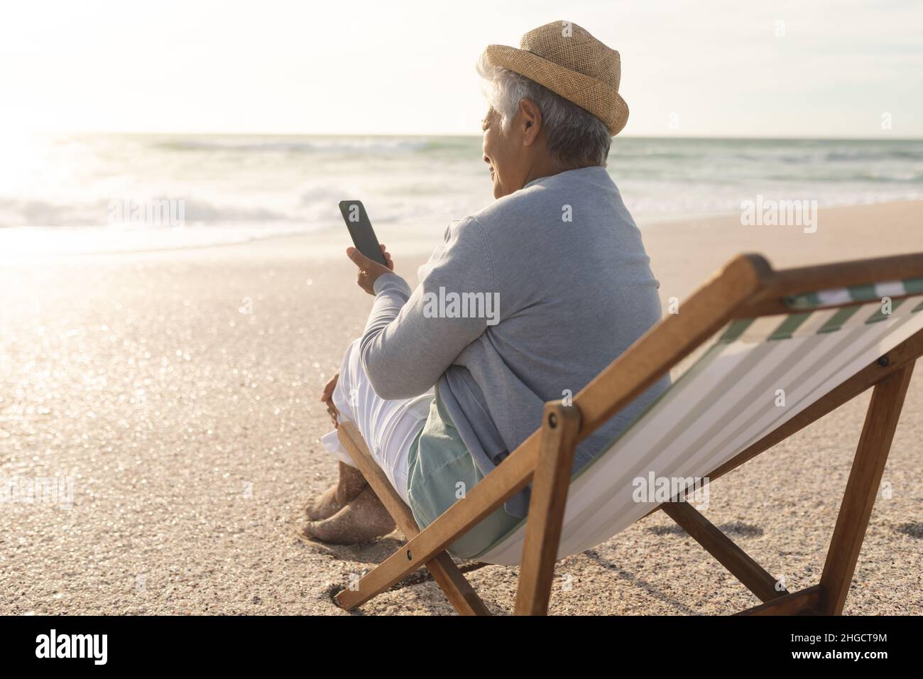 Ältere Frau aus der Birazialzeit, die während des Sonnenuntergangs auf einem Stuhl am Strand sitzt und ein Smartphone für Videoanruf verwendet Stockfoto