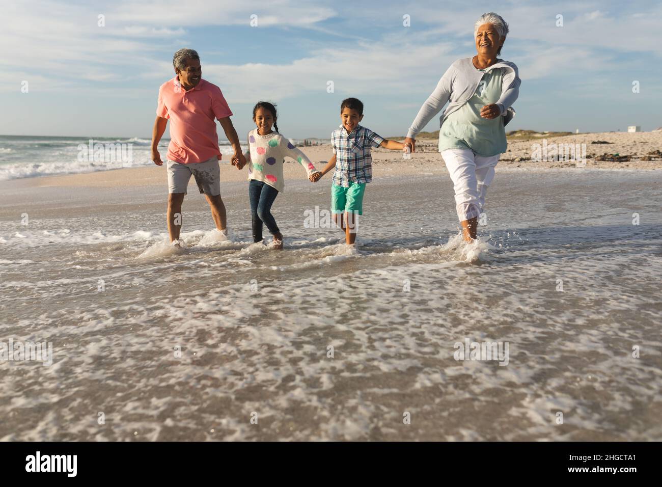 Fröhliche, multirassische Großeltern halten sich die Hände, während sie mit Enkelkindern am Strand an der Küste spazieren Stockfoto