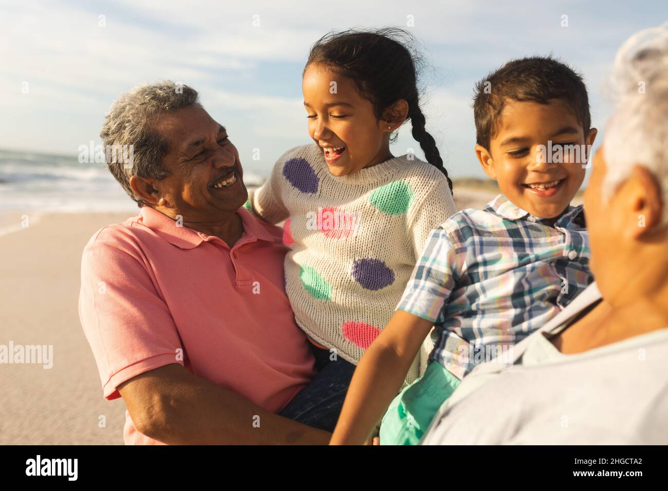 Lächelnde Großeltern aus verschiedenen Rassen, die Enkelkinder tragen, genießen das gemeinsame Wochenende am sonnigen Strand Stockfoto