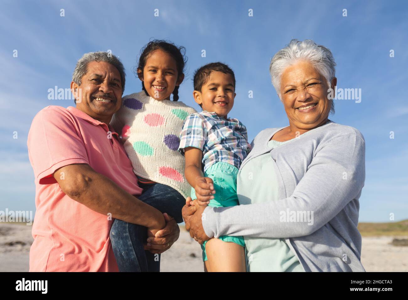 Porträt lächelnder Großeltern aus verschiedenen Rassen, die Enkel am Strand gegen den Himmel tragen Stockfoto