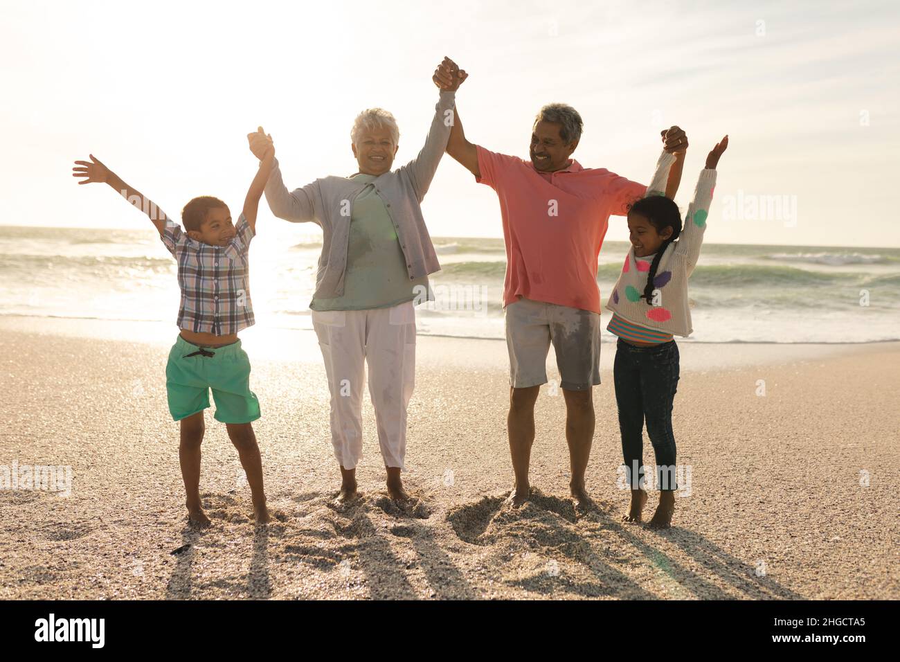 In voller Länge glückliche Großeltern und Enkelkinder, die am Strand die Hände gegen den Himmel halten Stockfoto