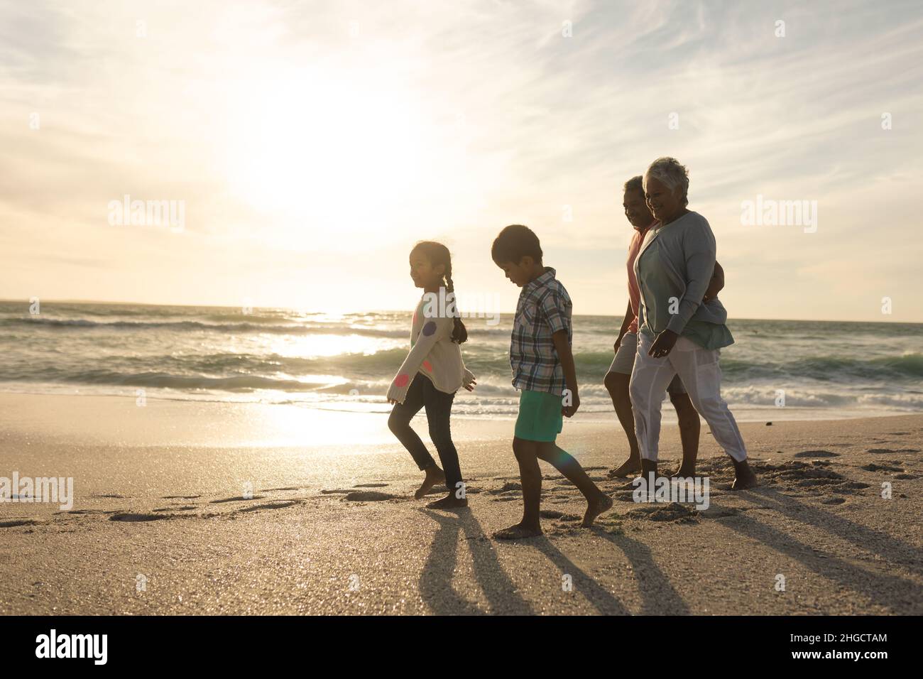 Seitenansicht der multirassischen Großeltern und Enkelkinder, die während des Sonnenuntergangs am Strand entlang gehen Stockfoto