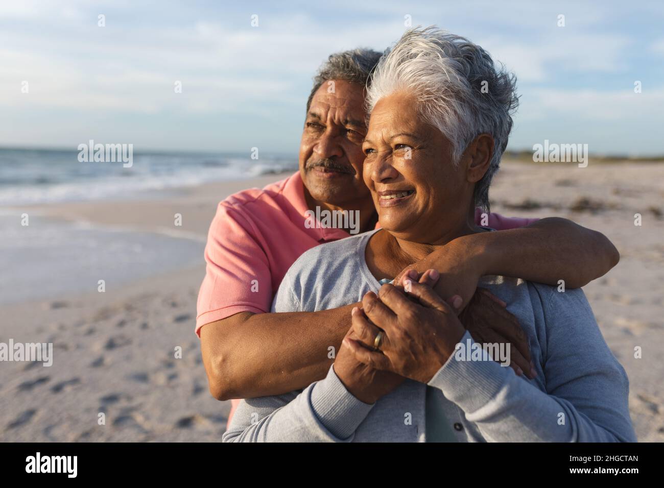 Nachdenkliches, mehrrassiges Paar, das sich umarmte, während es bei Sonnenuntergang auf den Strand schaute Stockfoto