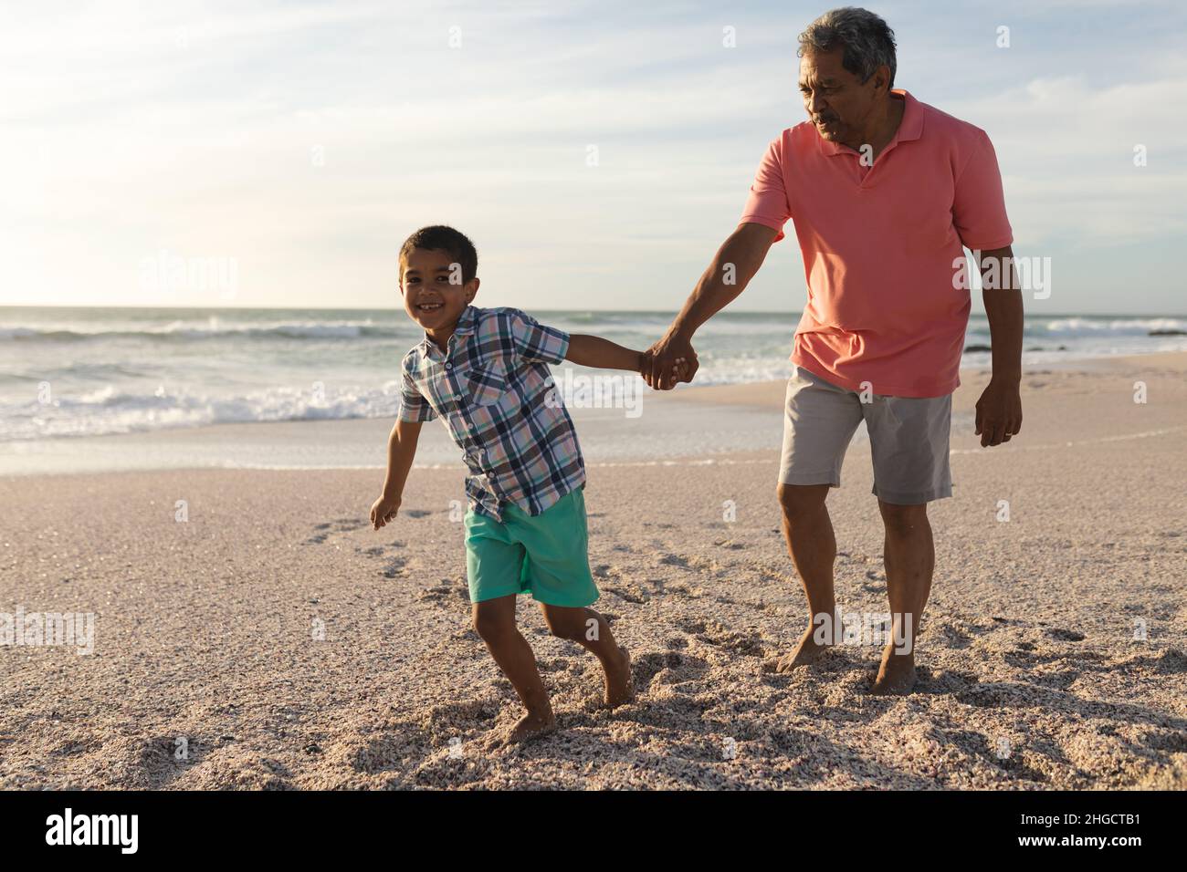 Lächelnder Biracial Junge mit der Hand des Großvaters, der am Strand gegen den Himmel läuft Stockfoto