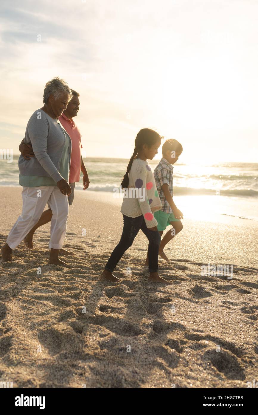 Seitenansicht der multirassischen Großeltern mit Arm um den Strand, der hinter Enkeln am Ufer herumläuft Stockfoto