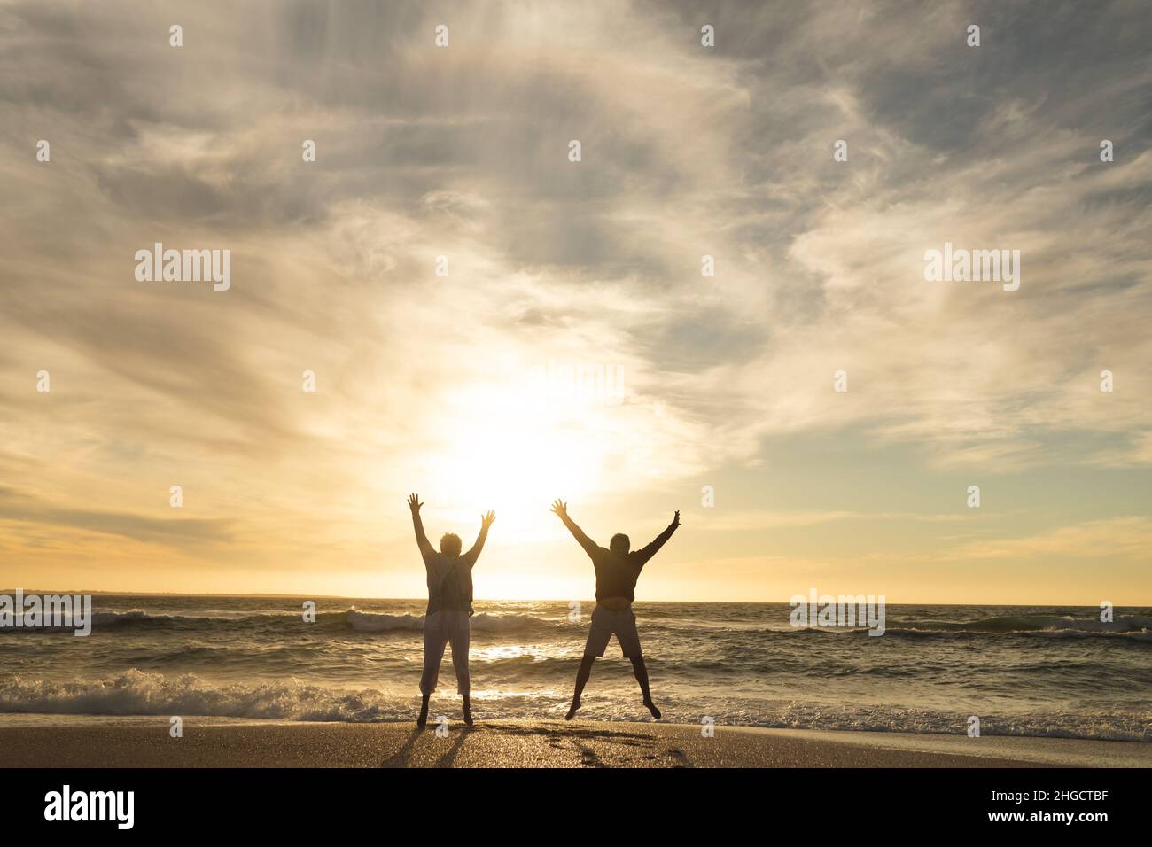 Sorgenfreies, multirassisches Seniorenpaar, das während des Sonnenuntergangs am Strand gegen den Himmel mit erhobenen Armen springt Stockfoto