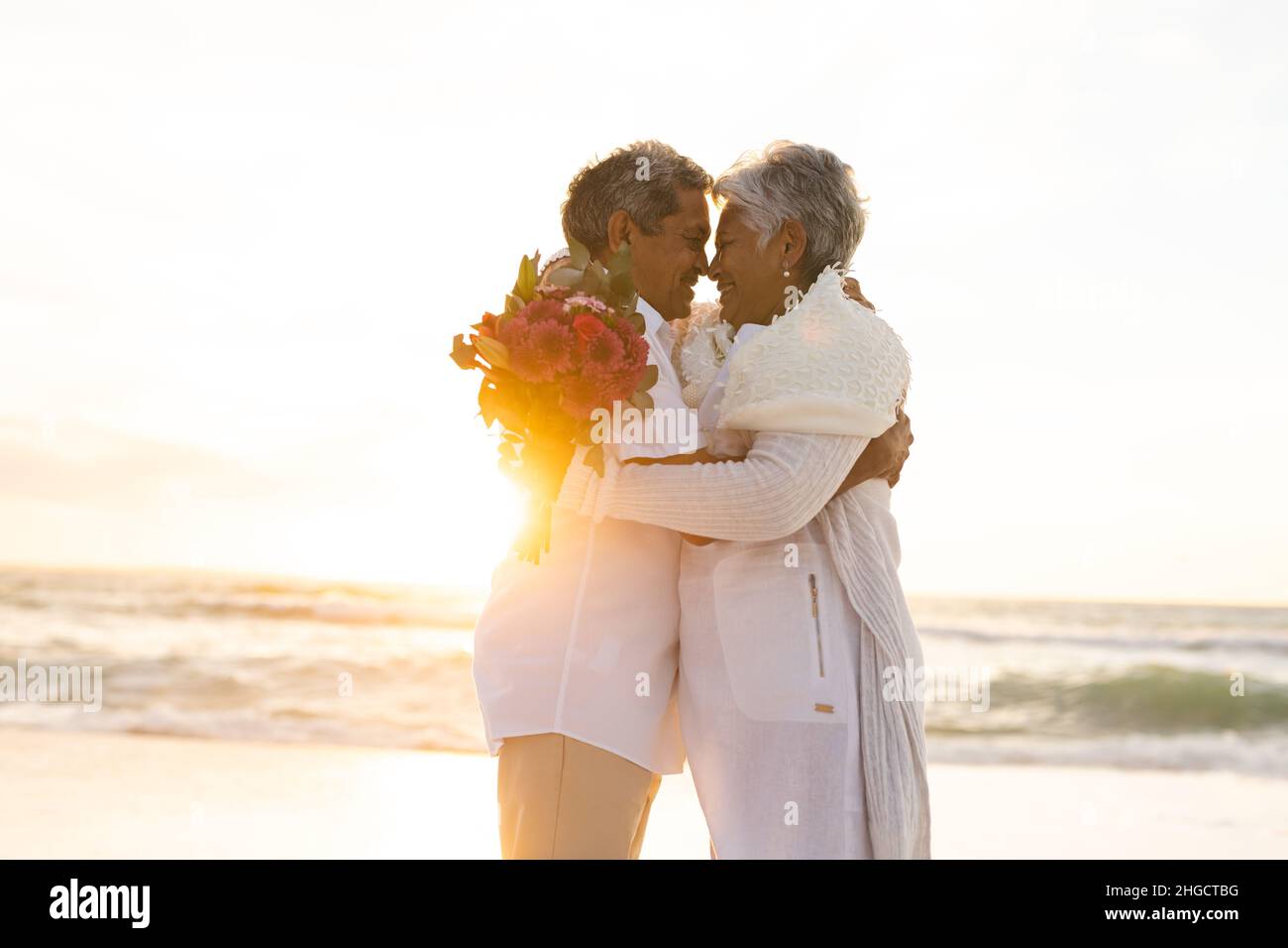 Ein glückliches, frisch verheiratetes, mehrrassiges Ehepaar umarmt sich, während es während des Sonnenuntergangs am Strand einen Blumenstrauß hält Stockfoto