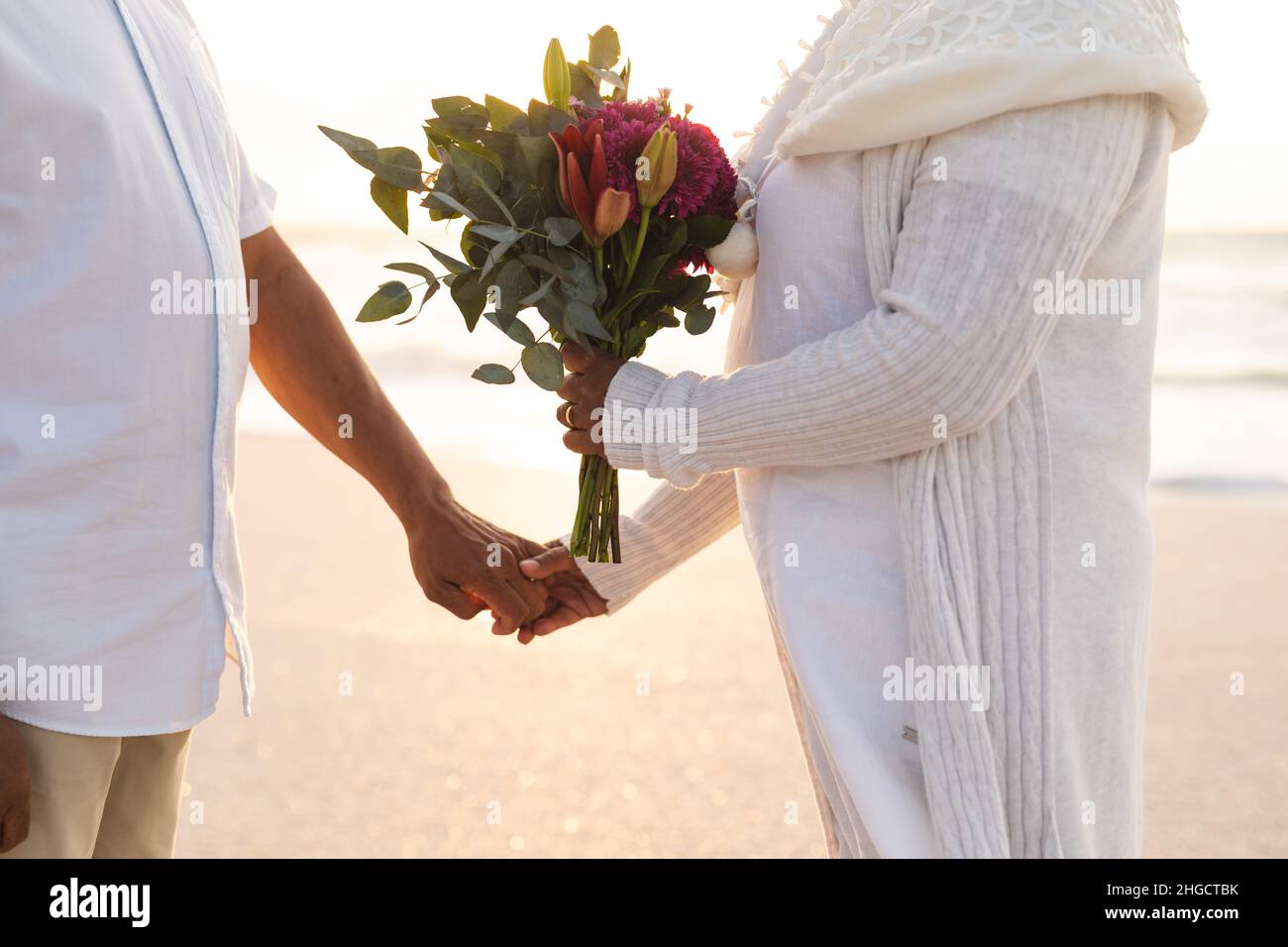 Mittelteil eines mehrrassischen älteren Paares, das während der Hochzeitszeremonie am Strand Hände mit Blumenstrauß hält Stockfoto