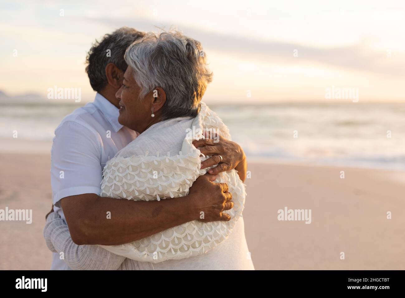 Glückliche, frisch vermählte, multirassische ältere Braut und Bräutigam umarmen sich am Strand während des Sonnenuntergangs Stockfoto
