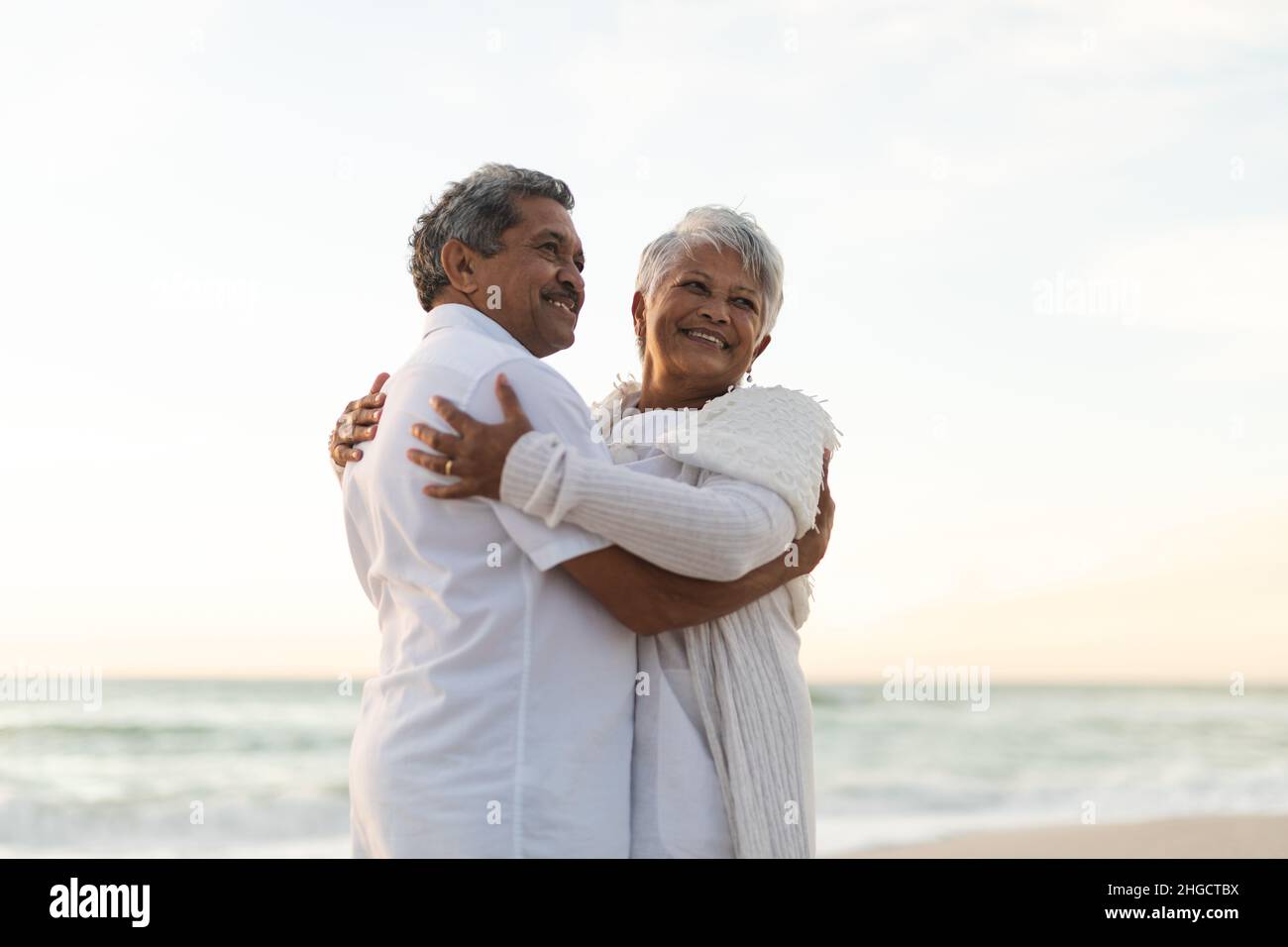 Lächelndes, frisch verheiratetes, multirassisches Paar, das sich umarmte, während es während des Sonnenuntergangs auf den Strand schaute Stockfoto