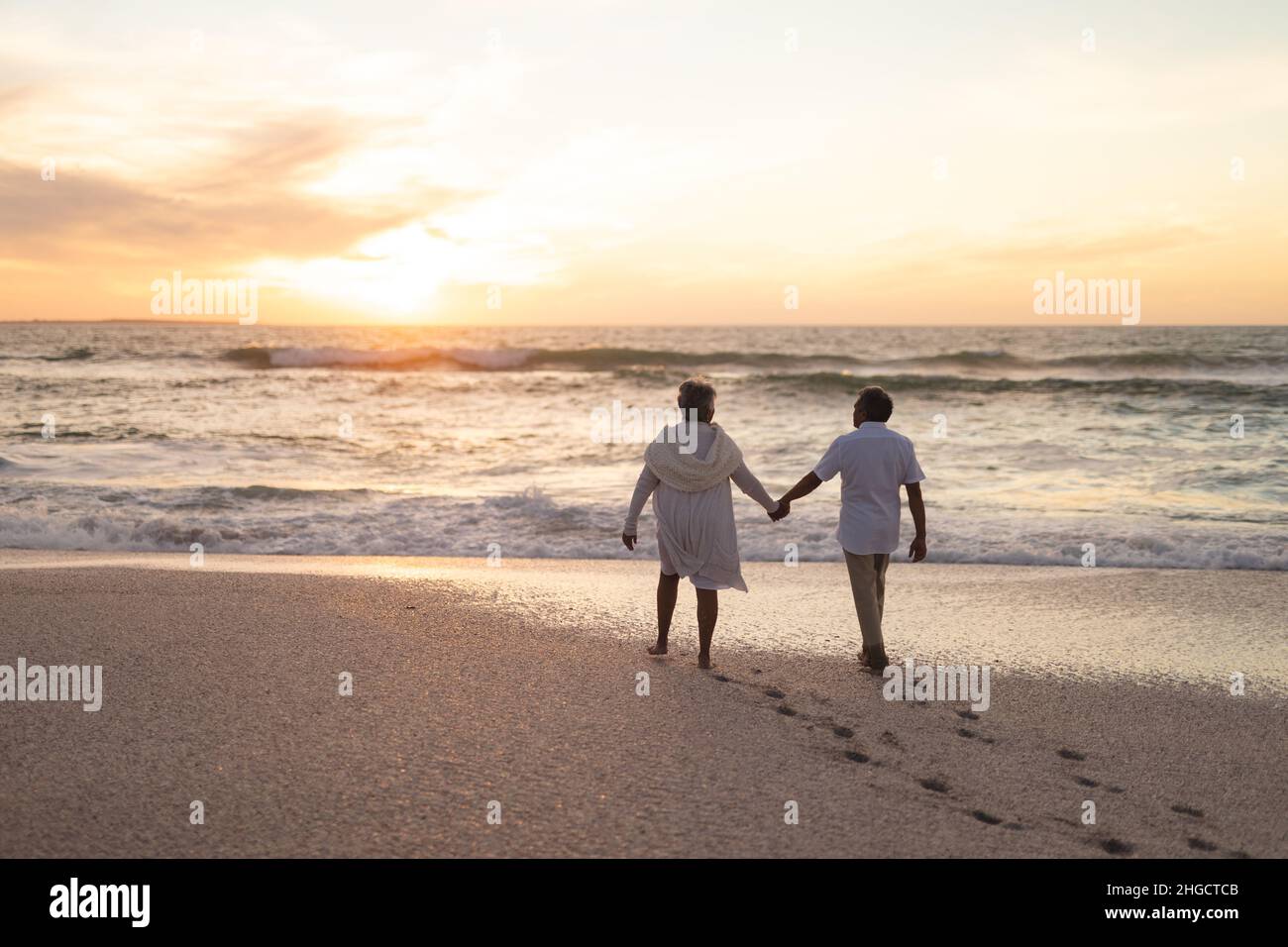 Rückansicht eines mehrrassischen Ehepaares, das während des Sonnenuntergangs am Strand herumläuft und die Hände hält Stockfoto