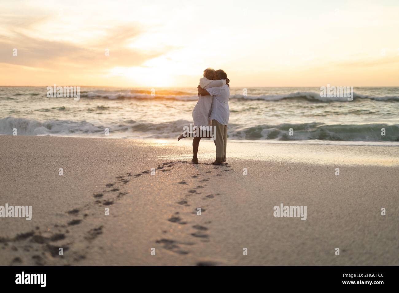 Seitenansicht des frisch vermählten mehrrassischen Ehepaares, das sich während des Sonnenuntergangs am Strand umarmt Stockfoto