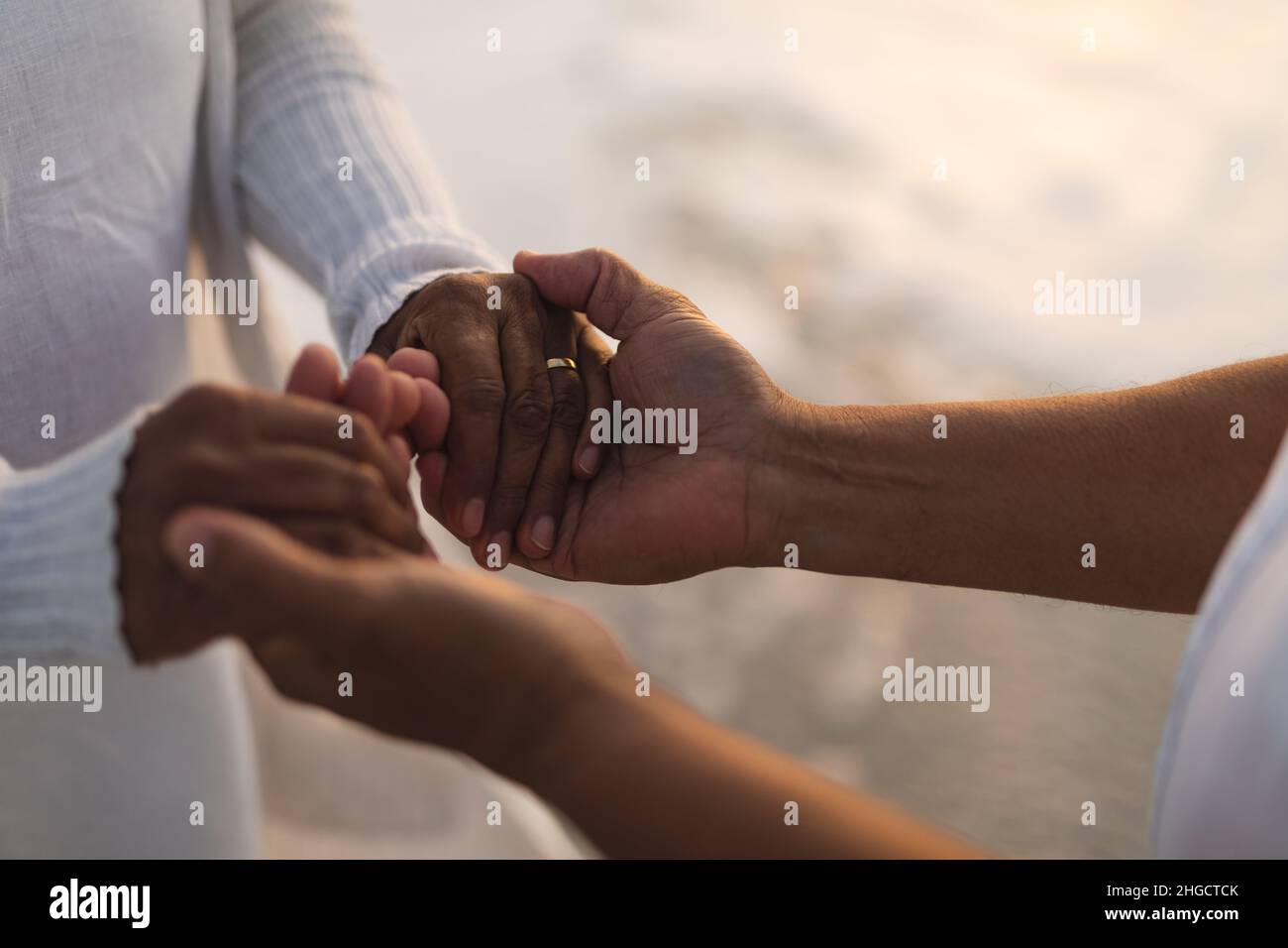 Mittelteil eines mehrrassigen, jungen Ehepaares, das während des Sonnenuntergangs am Strand die Hände hielt Stockfoto
