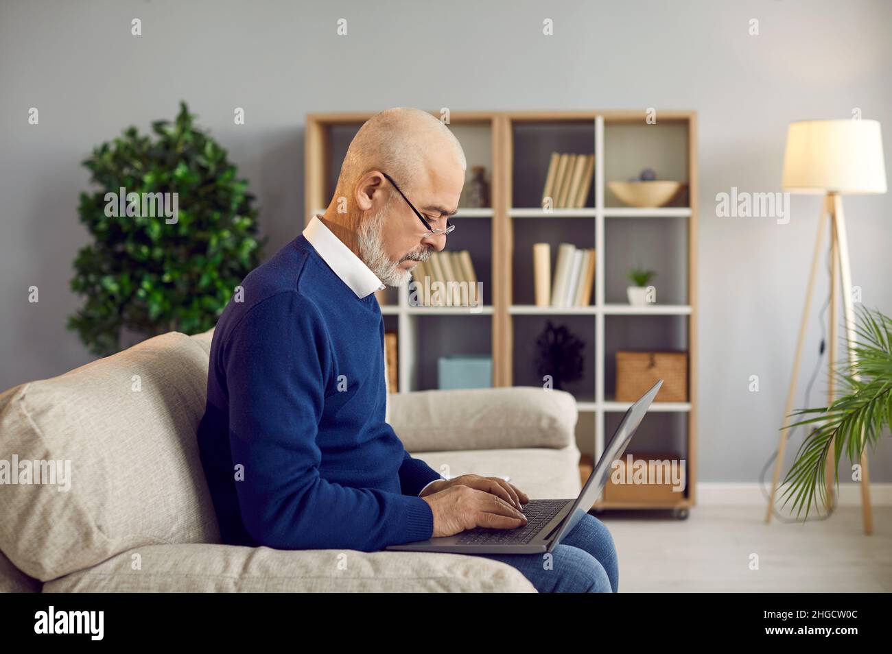 Seriös älterer Mann mit Brille sitzt auf dem Sofa zu Hause und arbeitet an seinem Laptop-Computer Stockfoto