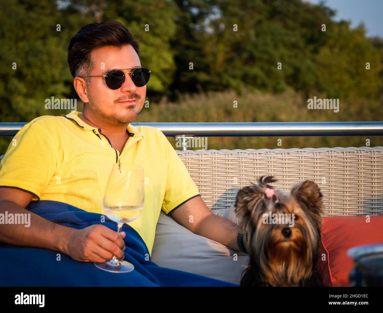 Junge schwarze Haaresbehaarung in legerer Kleidung und Sonnenbrille sitzt auf einer Terrasse, in eine Decke gehüllt, mit seinem yorkshire Terrier Hund und hält Glas Stockfoto