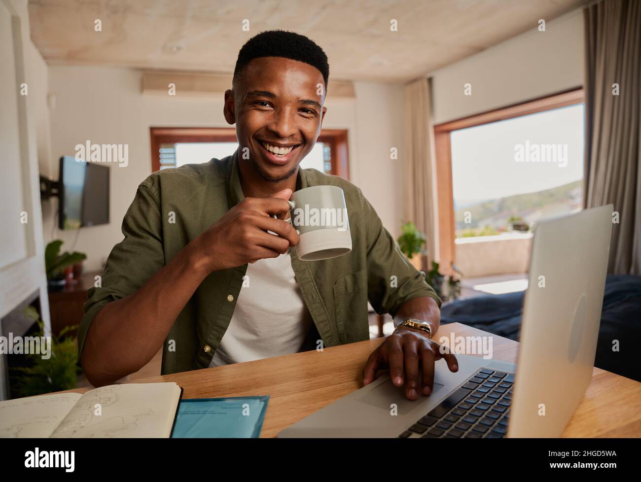 Porträt eines jungen erwachsenen schwarzen Mannes, der Becher hält und die Kamera anlächelt. Arbeiten aus der Ferne in einer modernen Wohnung. Stockfoto