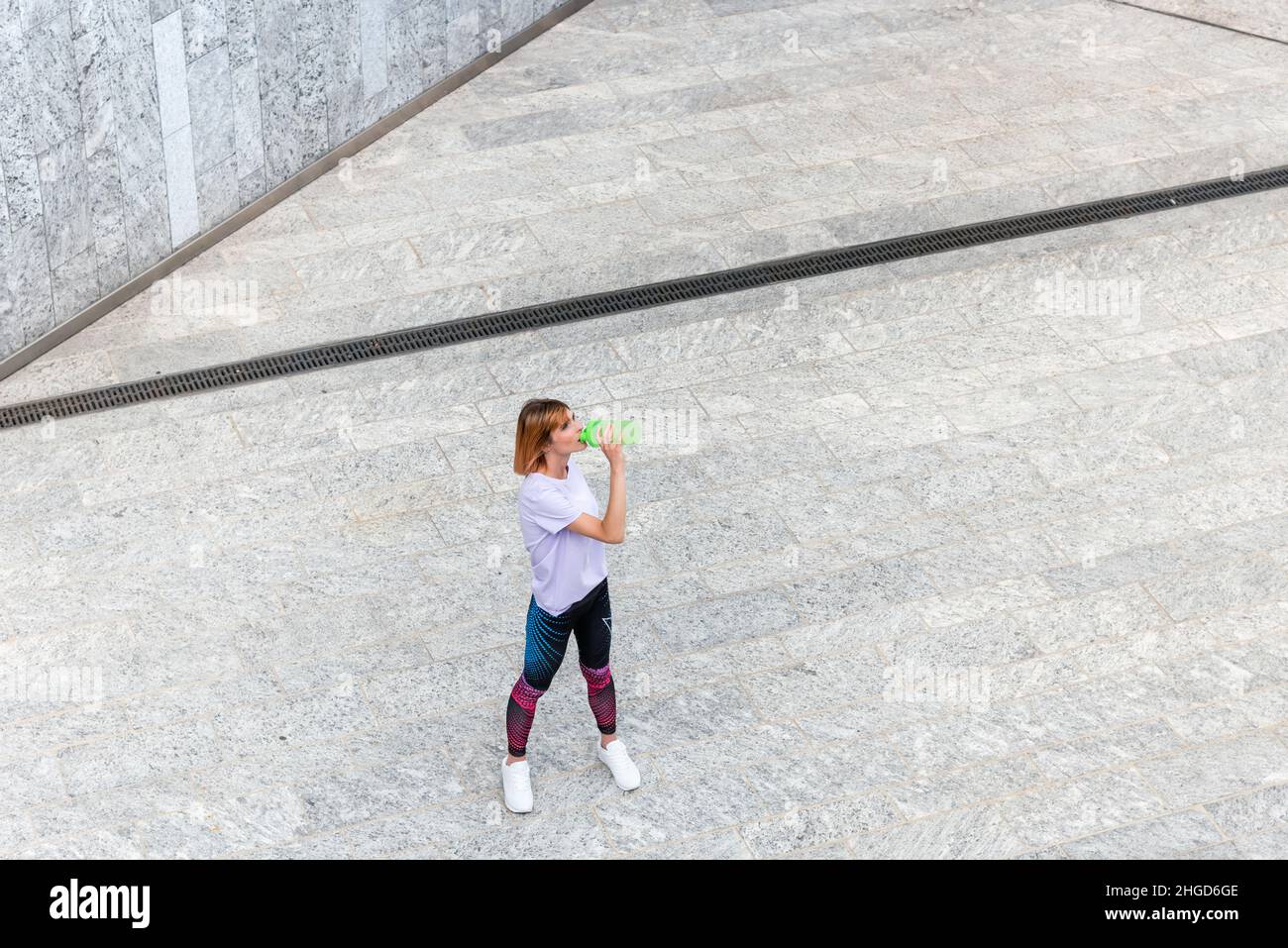 Von oben der jungen sportlichen Frau im aktiven Ohr, die erfrischendes Wasser aus der Flasche trinkt, während sie sich nach dem Training im Freien auf dem städtischen Platz ausruhte Stockfoto