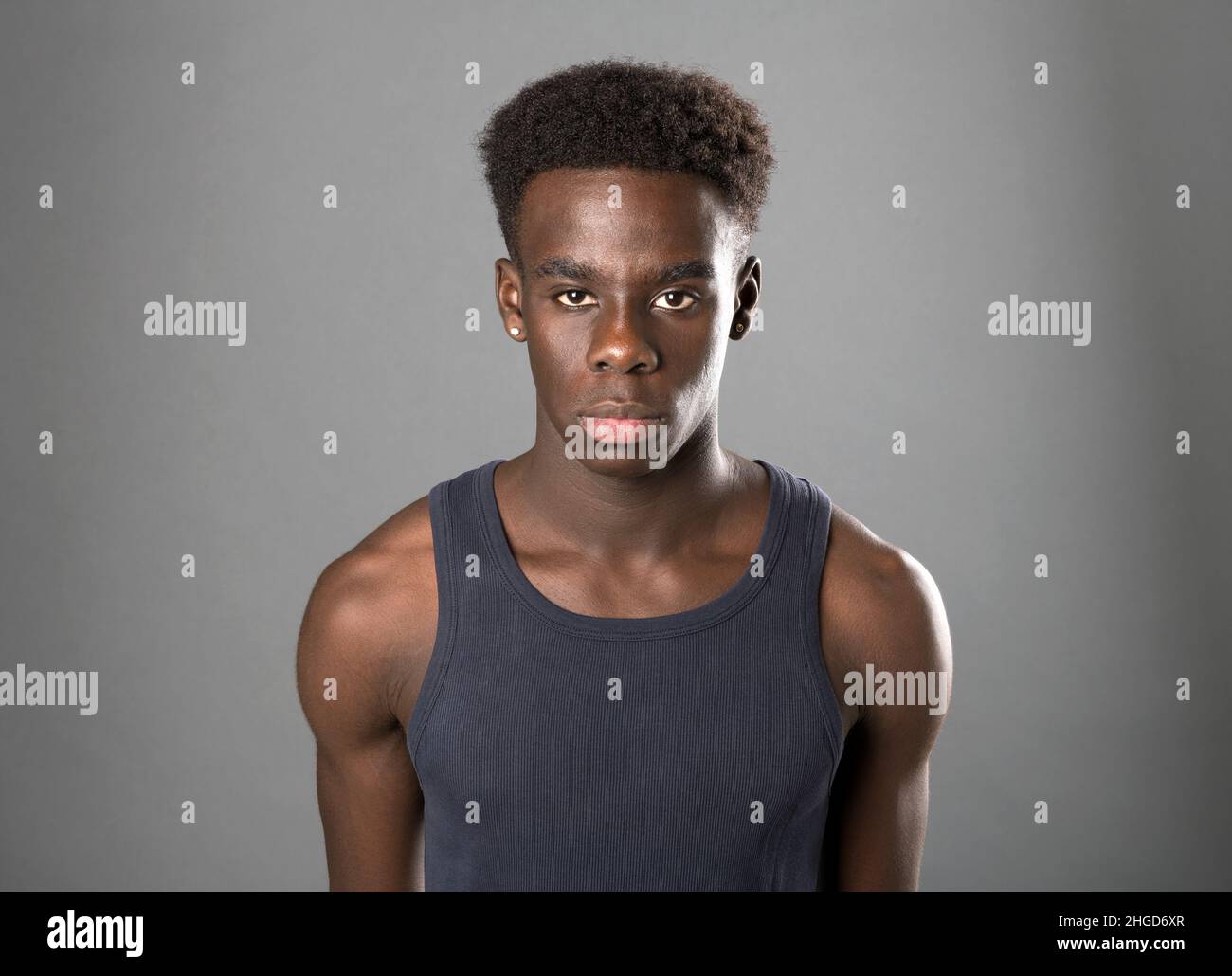 Selbstbewusster, muskulöser afroamerikanischer Mann im grauen Tank-T-Shirt, der im Studio mit ernstem Gesicht auf grauem Hintergrund auf die Kamera schaut Stockfoto