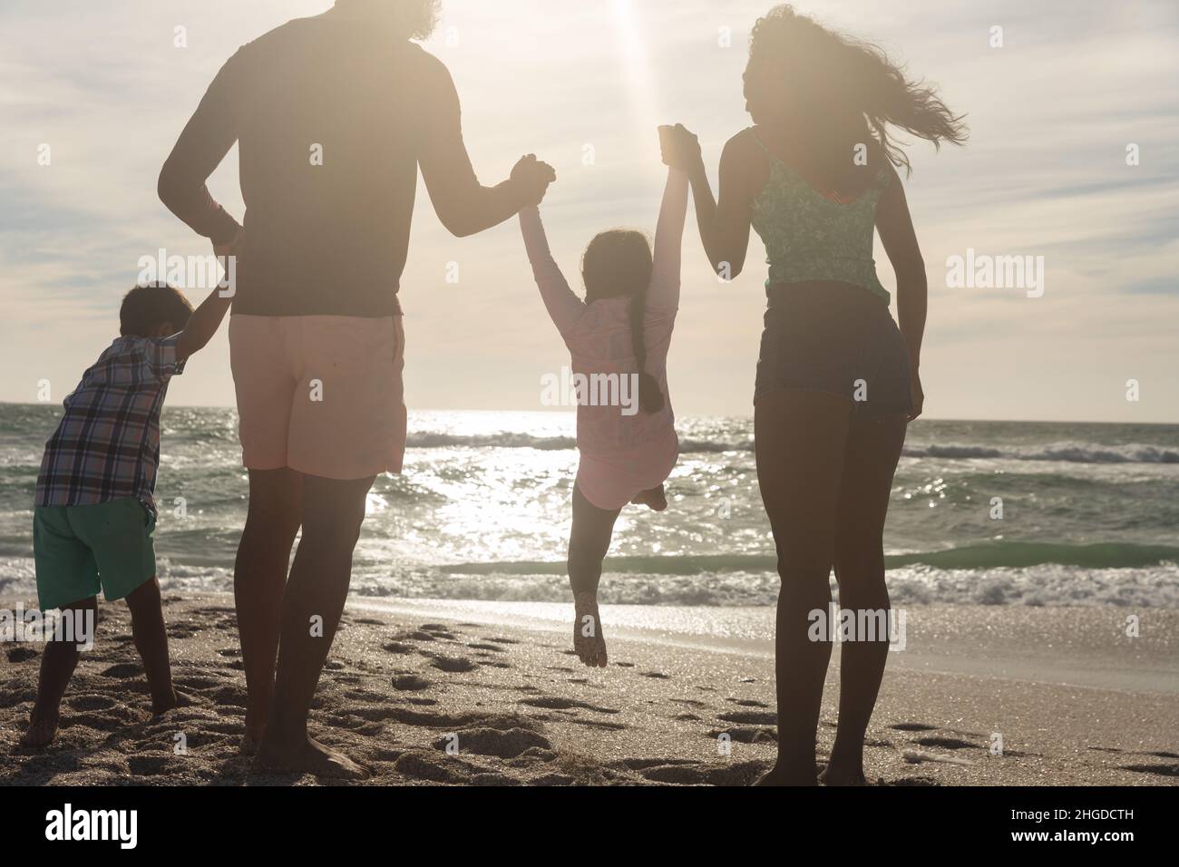 Rückansicht einer verspielten multirassischen Familie, die sich die Hände hält und einen sonnigen Tag am Strand genießt Stockfoto