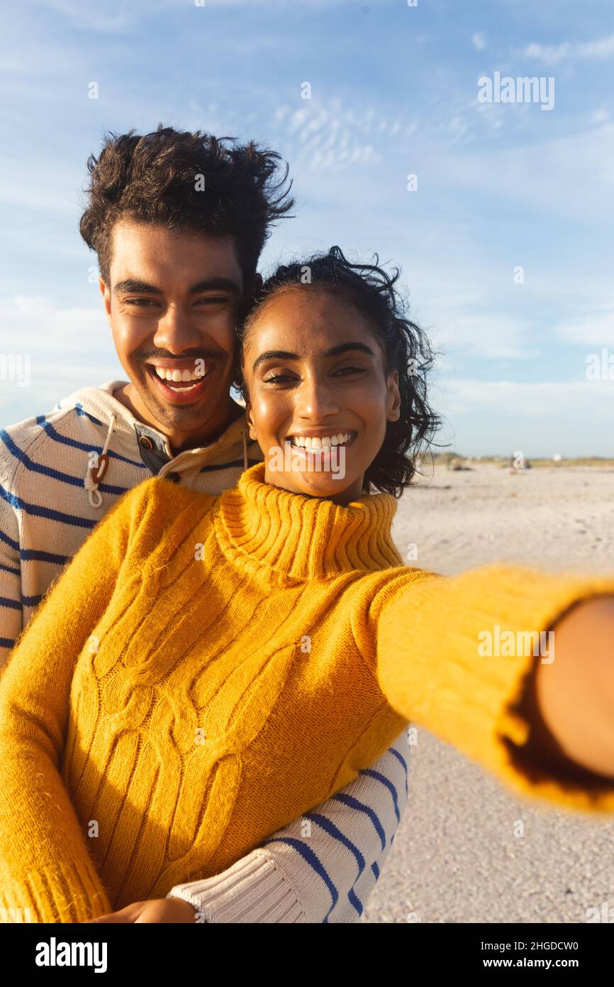 Porträt eines fröhlichen multirassischen Paares, das am sonnigen Tag am Strand Selfie gegen den Himmel nimmt Stockfoto