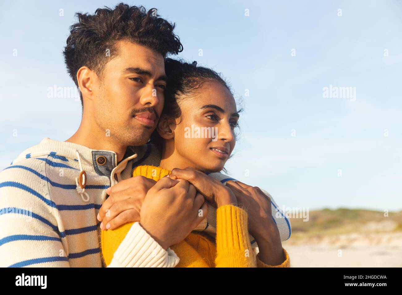 Nachdenkliches multirassisches Paar, das sich umarmt, während es am sonnigen Tag auf den Strand gegen den Himmel blickt Stockfoto