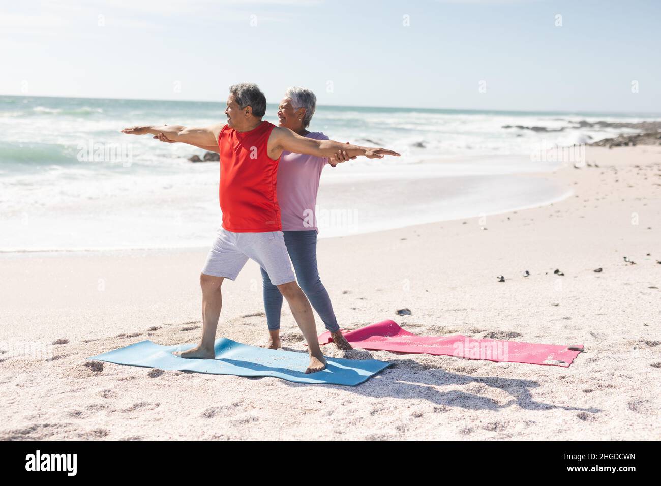 Ältere Birazialfrau hilft Mann mit Krieger Yoga Haltung üben am Strand gegen Himmel Stockfoto