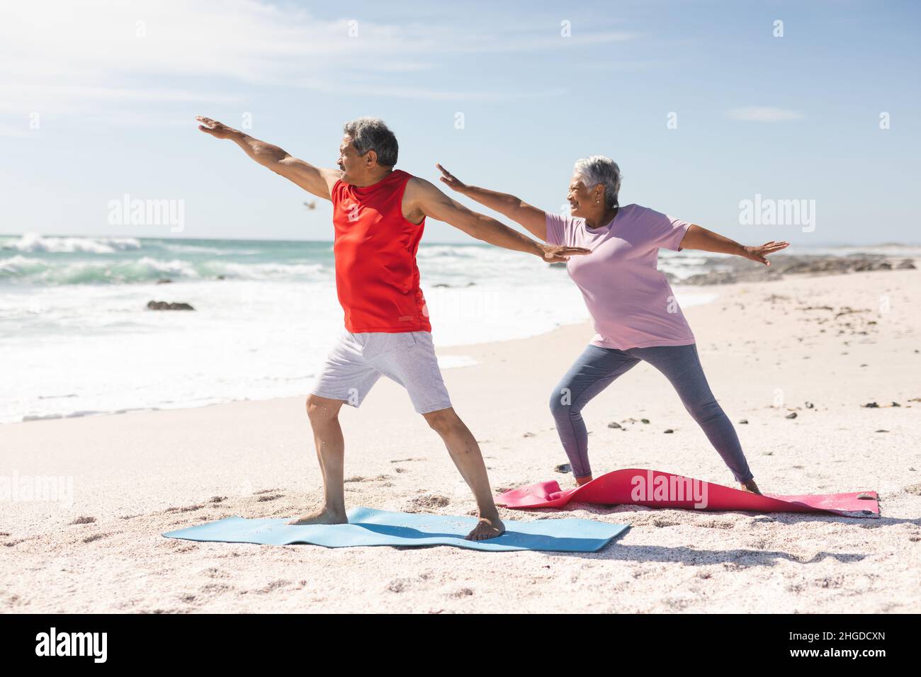 Biracial Senior Pärchen üben Krieger Yoga auf Matten am Strand gegen Himmel während sonnigen Tag Stockfoto
