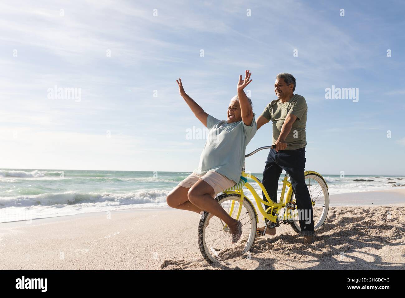 Fröhliches mehrrassiges Seniorenpaar, das am sonnigen Tag mit dem Fahrrad am Strand in den Ruhestand geht Stockfoto