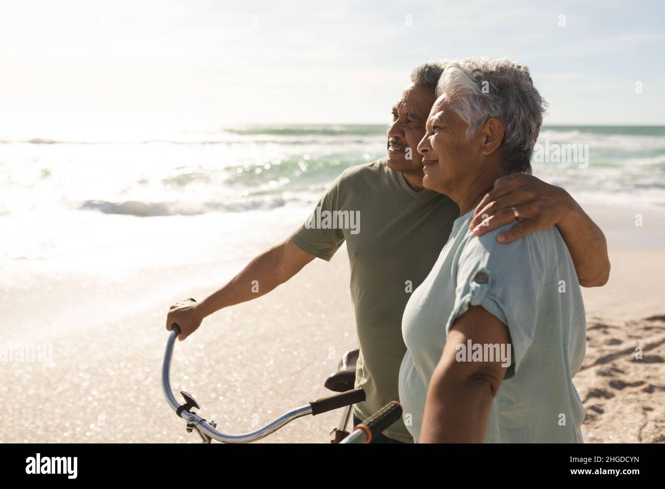 Nachdenkliches, multirassisches Seniorenpaar blickt weg, während es mit Fahrrädern am sonnigen Strand steht Stockfoto