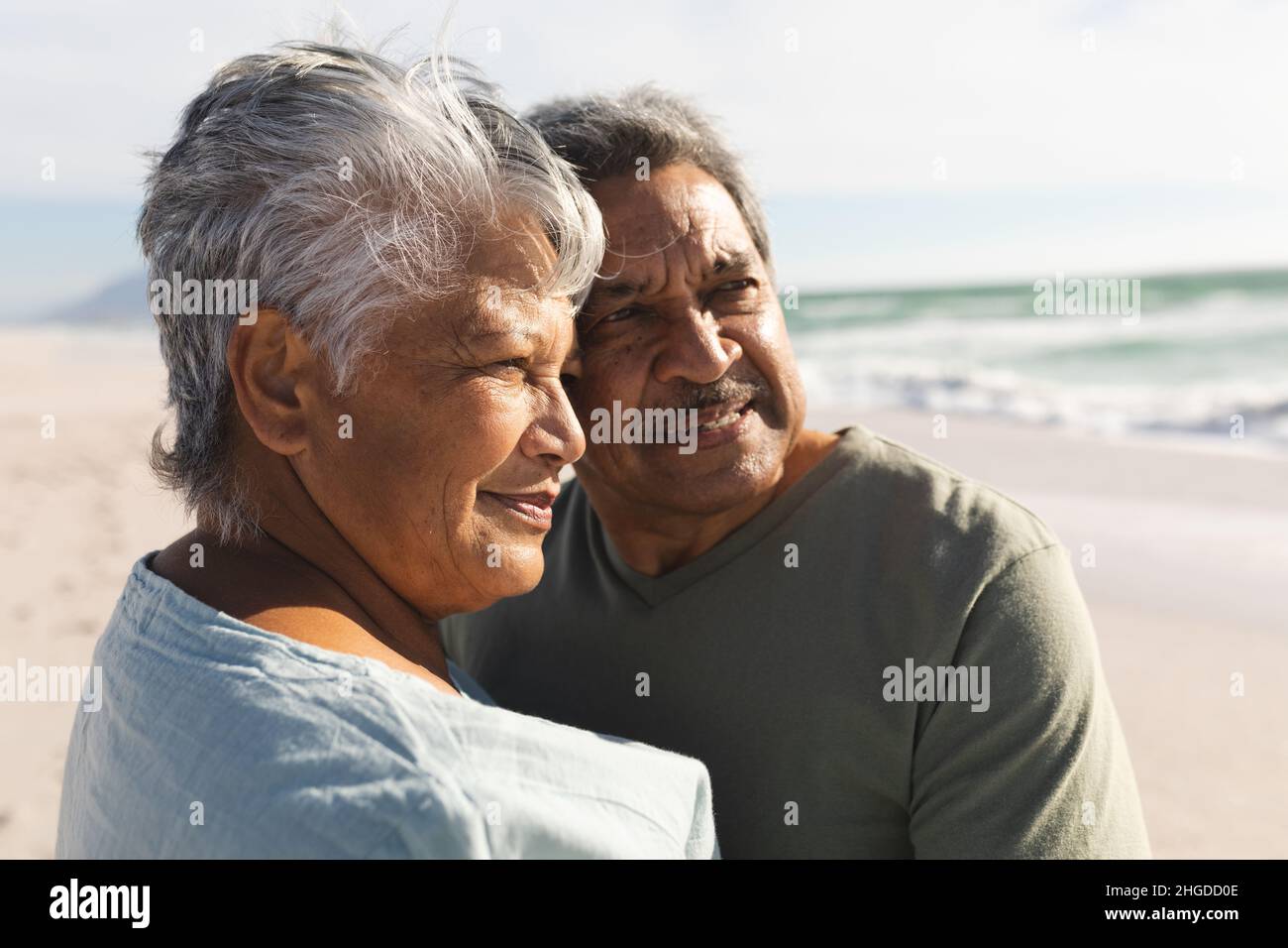 Nachdenkliches mehrrassiges Senioren-Paar, das an einem sonnigen Tag am Strand steht und zusammen wegschaut Stockfoto