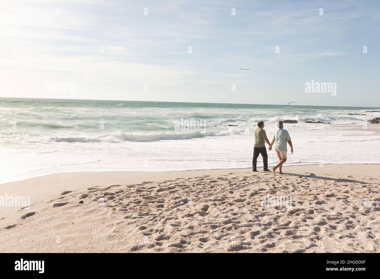 Die ganze Länge eines mehrrassischen Seniorenpaares, das am sonnigen Strand die Hände am Ufer hält Stockfoto