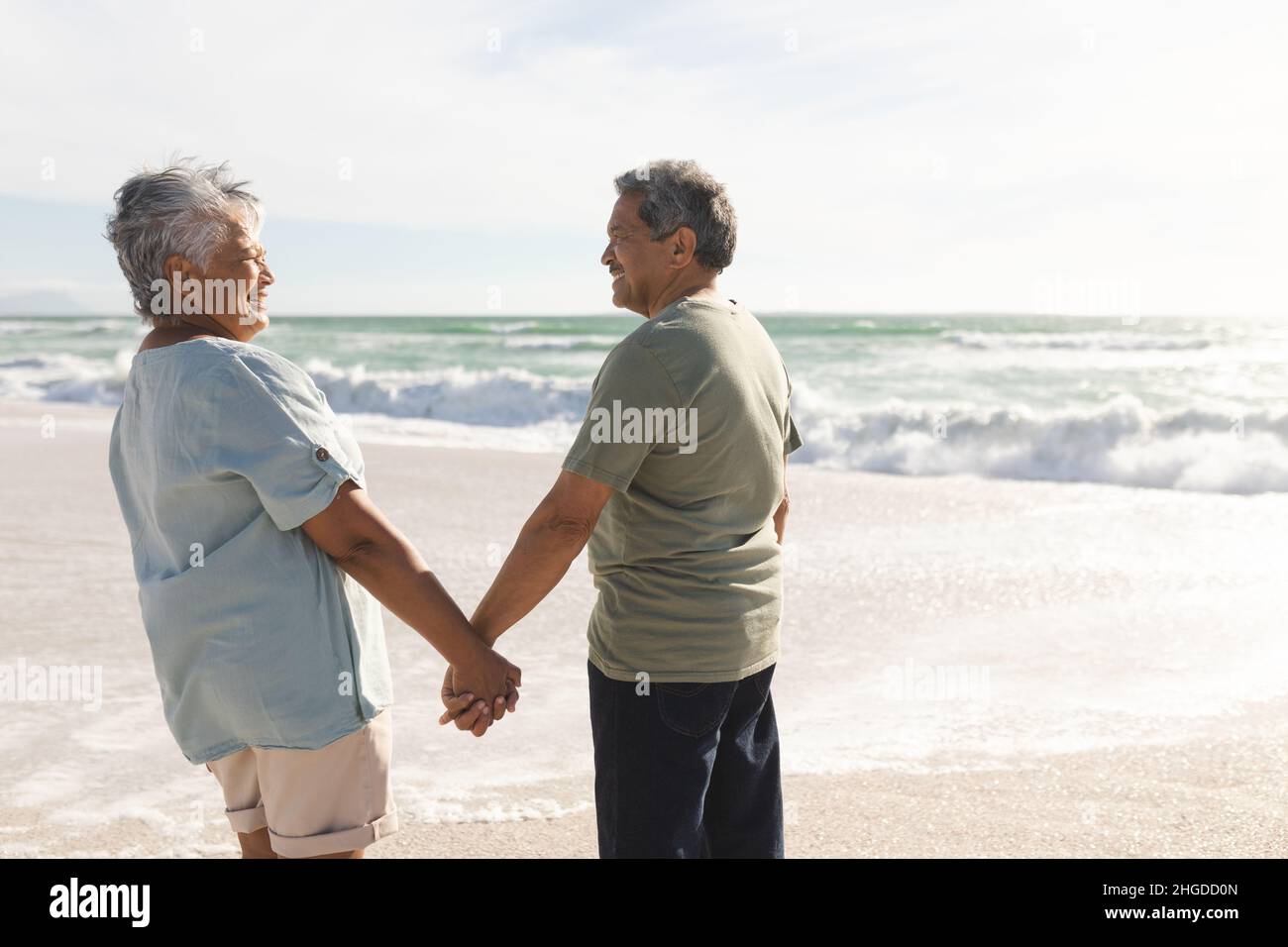 Glückliches, mehrrassiges Senioren-Paar im Ruhestand, das sich am sonnigen Strand die Hände hielt und sich ansah Stockfoto