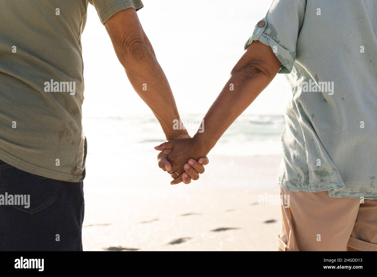 Mittlere Rückansicht eines mehrrassigen älteren Ehepaares, das an einem sonnigen Tag die Hände am Strand hielt Stockfoto