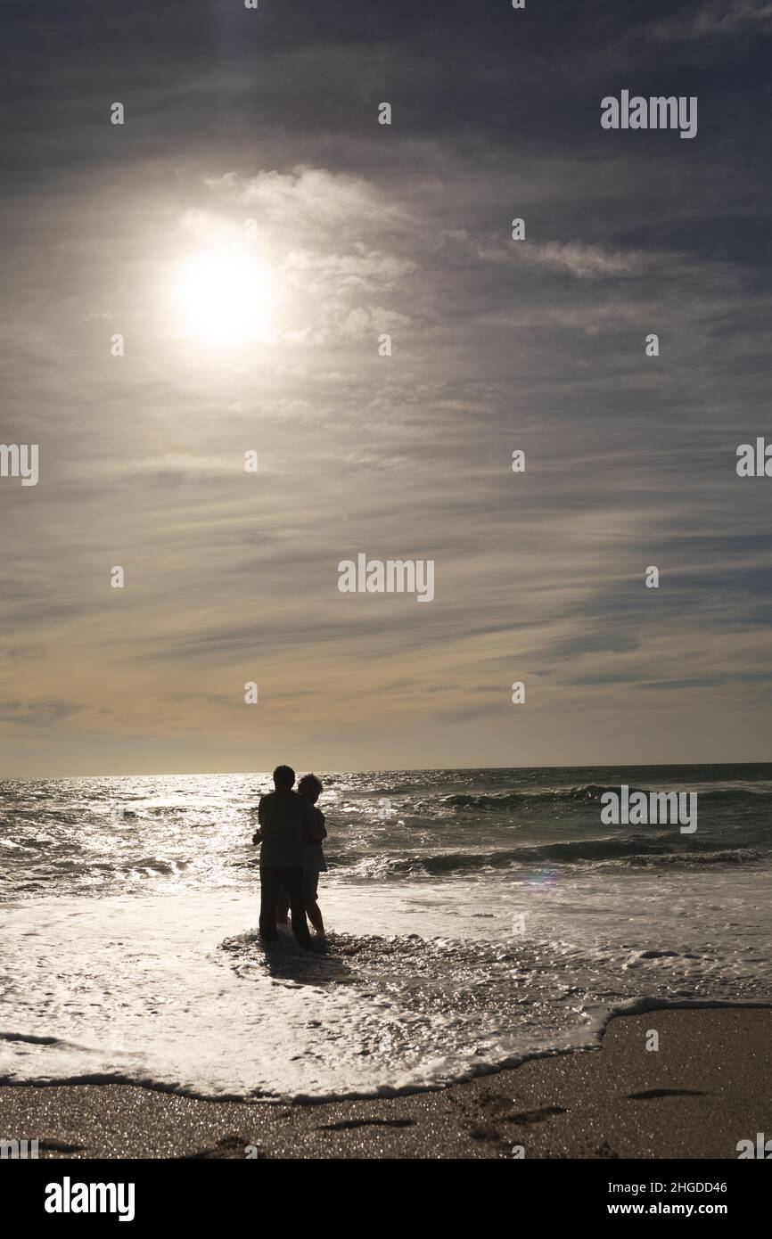 Ältere pensionierte multirassische Paare genießen Wellen zusammen am Ufer am Strand gegen den Himmel Stockfoto