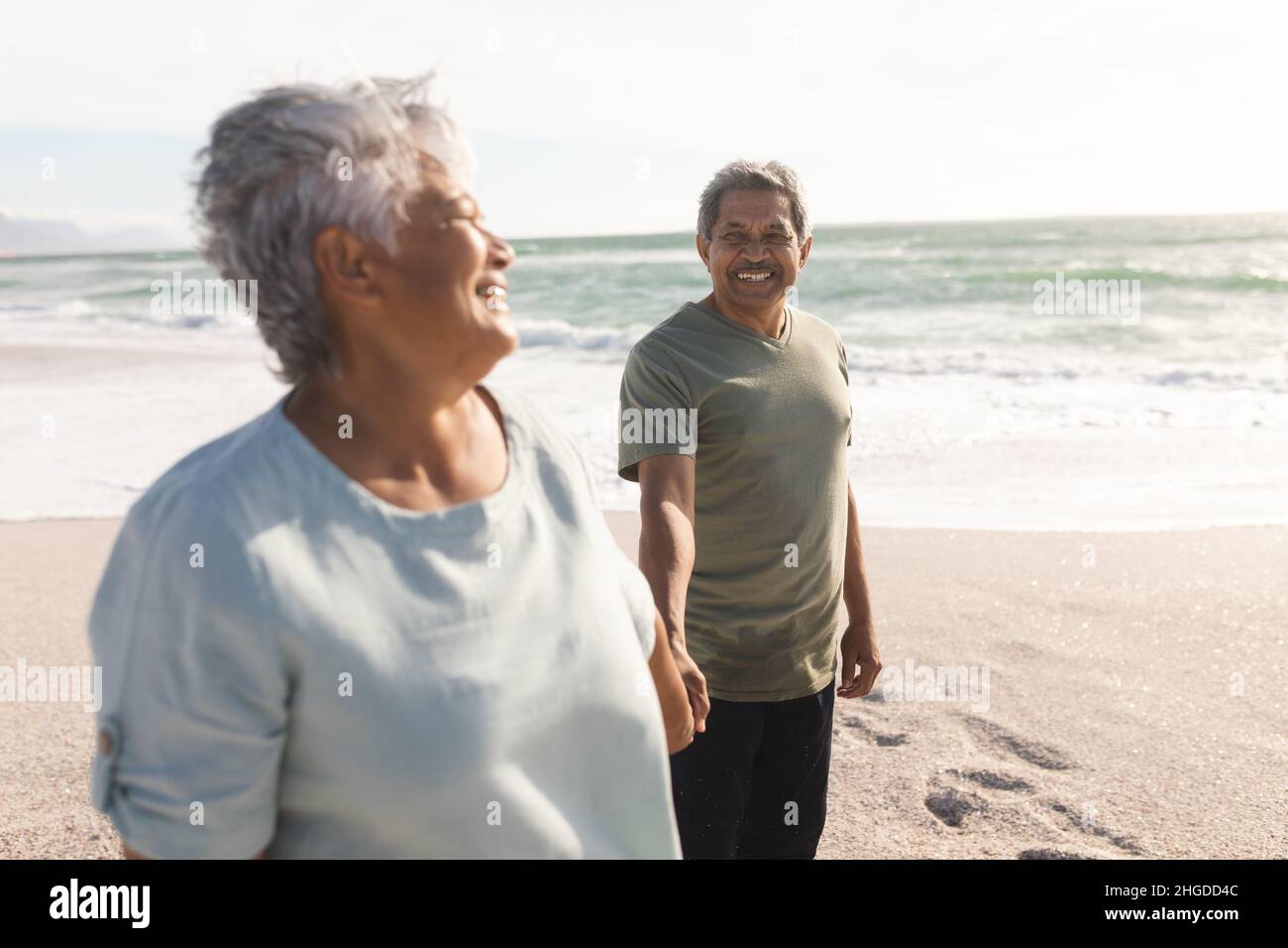 Ein lächelndes, mehrrassiges Paar hält sich die Hände, während es einen sonnigen Tag am Strand genießt Stockfoto