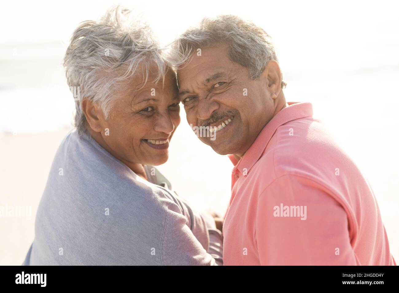 Porträt eines lächelnden, multirassischen Seniorenpaares, das über die Schultern blickt und am sonnigen Strand sitzt Stockfoto