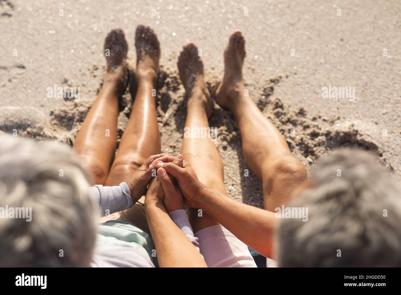 Blick aus dem hohen Winkel auf ein liebevolles, multirassisches Seniorenpaar, das die Hände am sonnigen Strand auf Sand hält Stockfoto