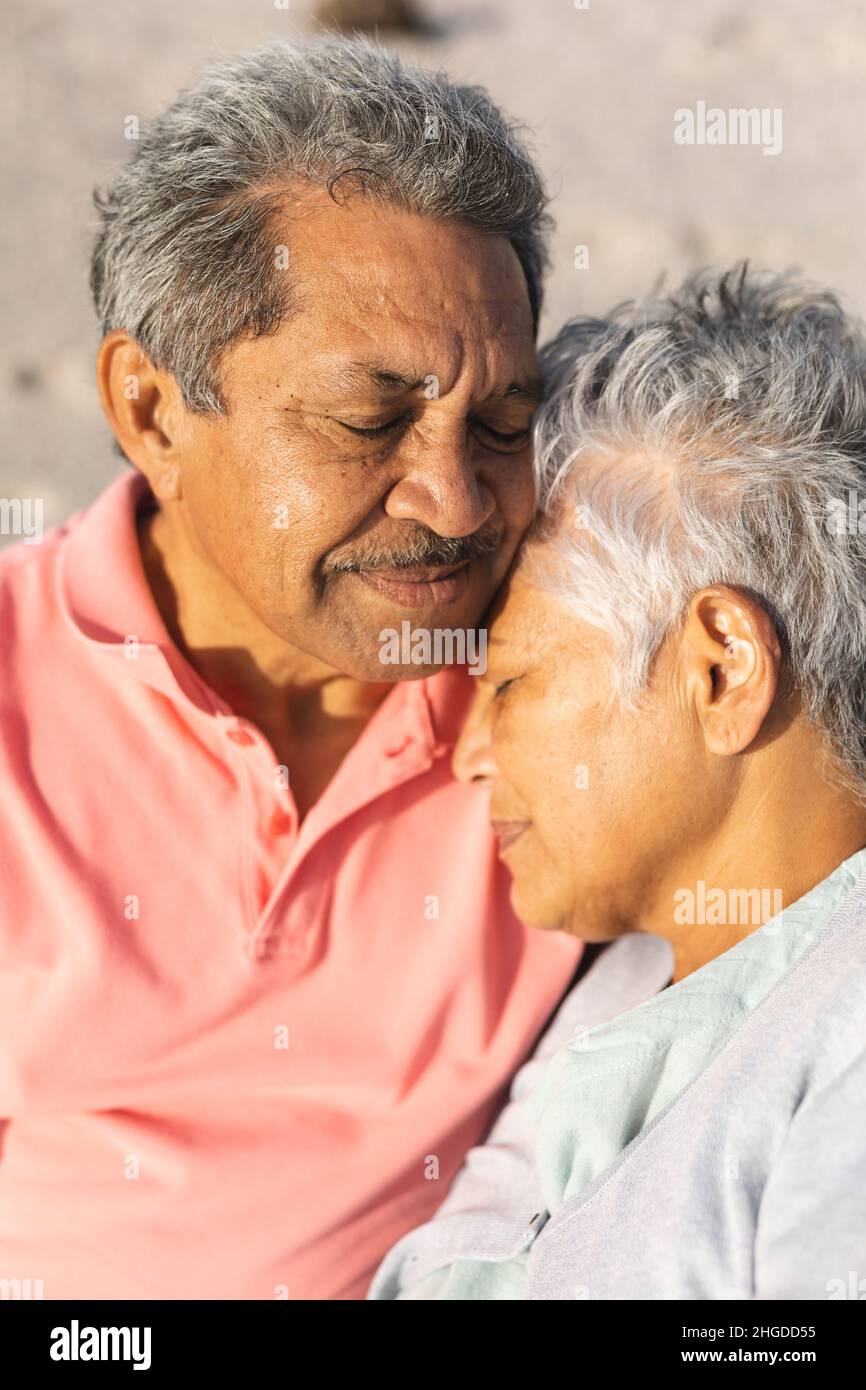 Romantisches mehrrassisches Senioren-Paar, das an einem sonnigen Tag mit geschlossenen Augen am Strand sitzt Stockfoto