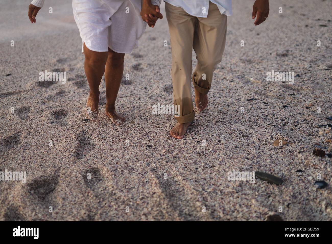 Niedriger Teil eines älteren multirassischen Paares, das die Hände hält und barfuß auf Sand am Strand läuft Stockfoto