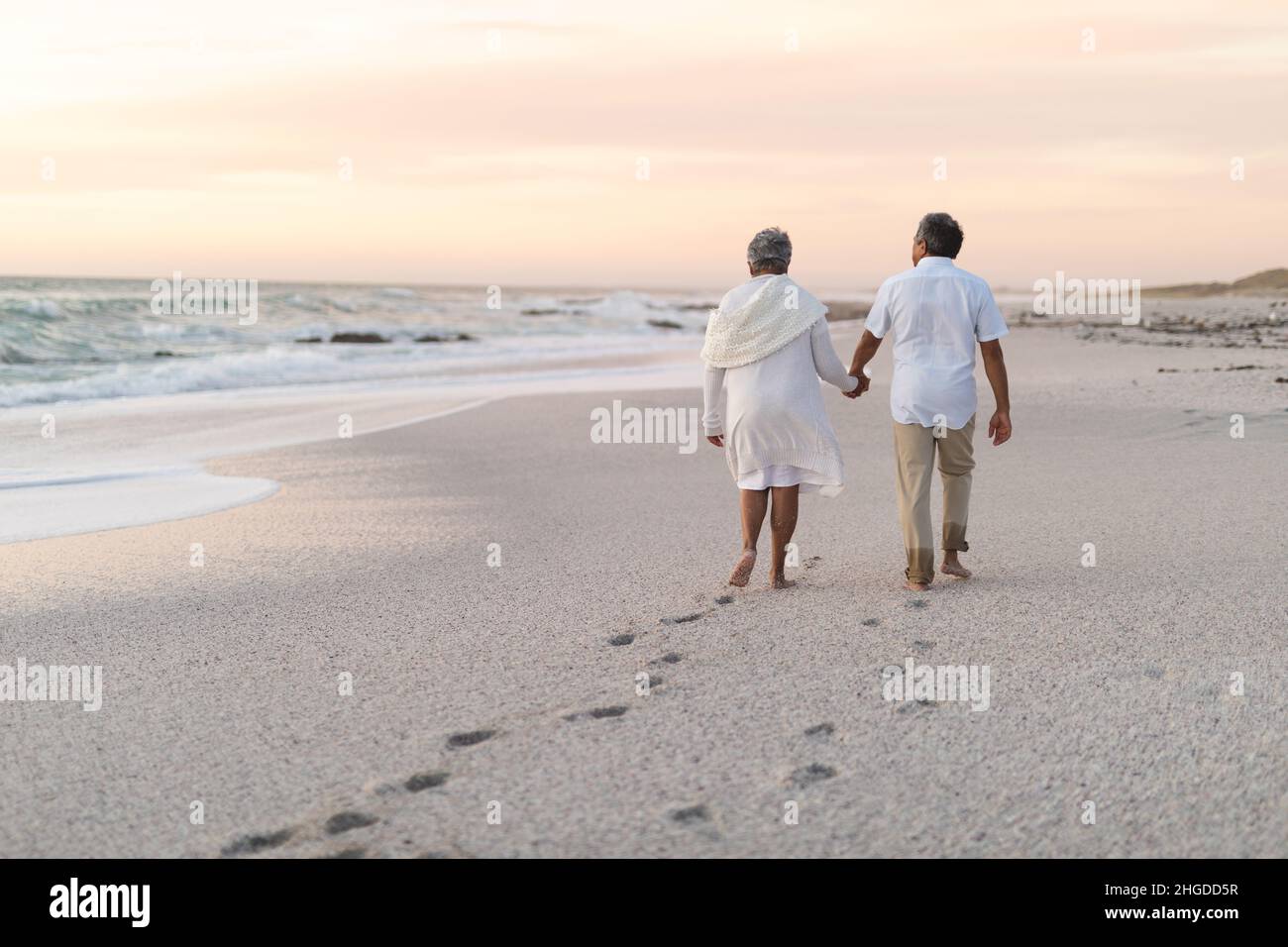 Multirassisches Seniorenpaar hält die Hände zusammen und hinterlässt Fußabdrücke auf Sand Stockfoto