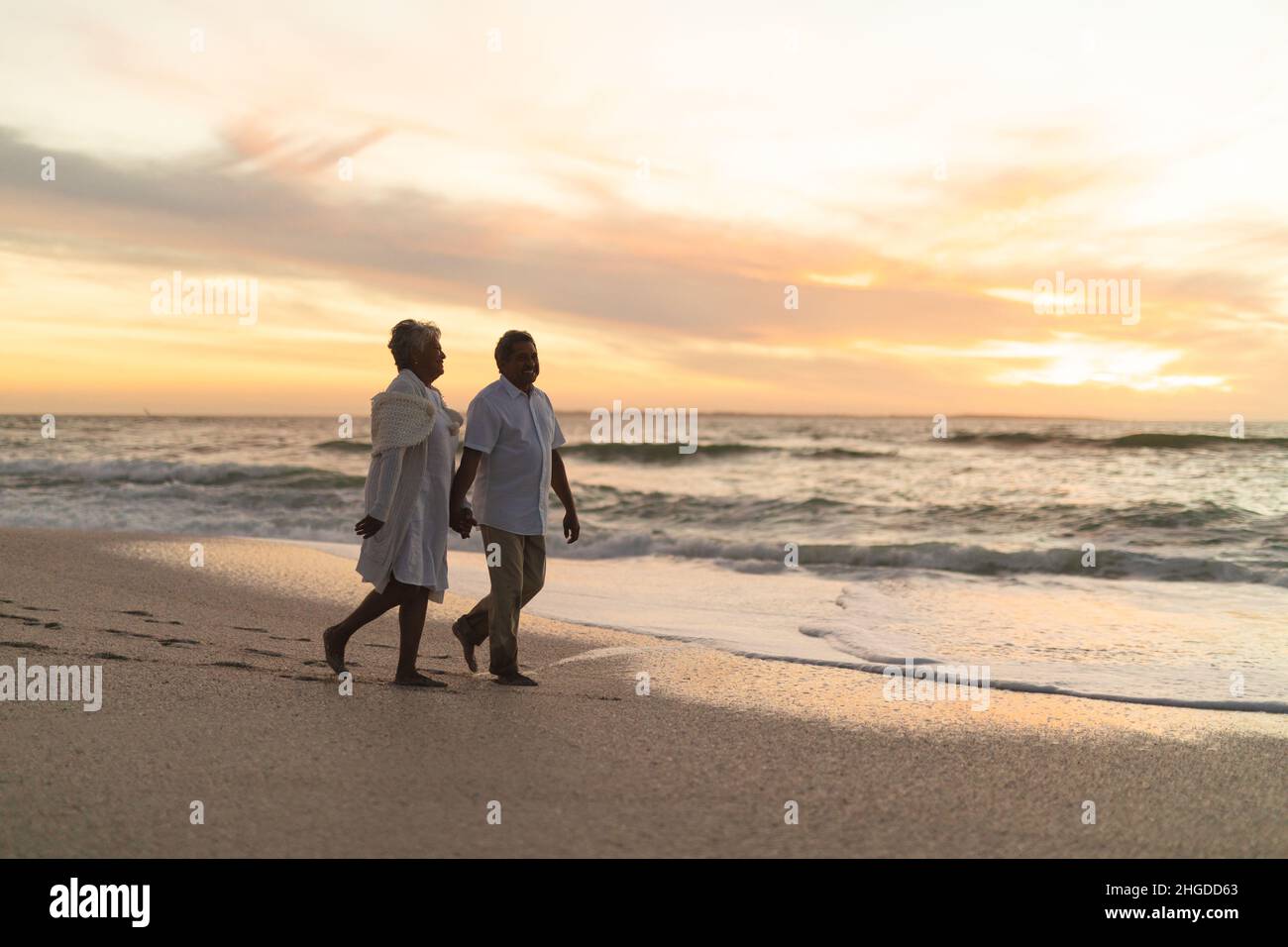 Die gesamte Länge eines mehrrassischen Seniorenpaares, das gemeinsam den Sonnenuntergang verbringt, während es am Strand gegen den Himmel läuft Stockfoto