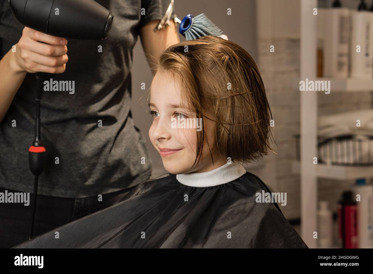 Friseur Föhnen Modell kurze nasse Haare nach dem Schneiden. Das zufriedene Kind sieht im Spiegel aus und bewundert das neue Image. Das kurzhaarige Mädchen lächelt im Friseurladen Stockfoto