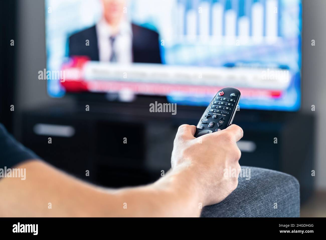 Mann, der Fernsehnachrichten anschaut und auf der Couch sitzt. Fernbedienung des Fernsehers in der Hand. Netzwerkkanal für Politik, Wirtschaft oder Finanzen. Stockfoto